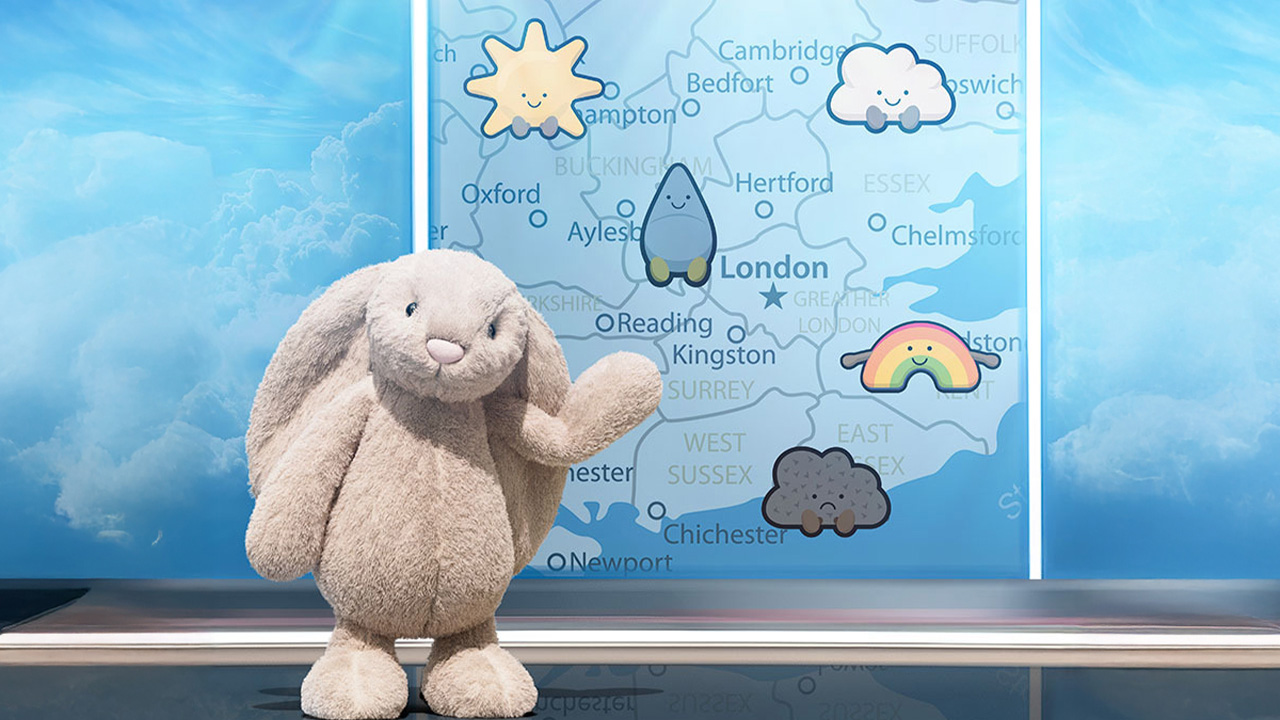 A Jellycat plush bunny stands in front of a whimsical weather map featuring cute cloud and rainbow illustrations.