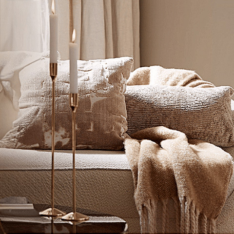 Cozy living room with a plush sofa, textured pillows, a soft throw blanket, and lit candlesticks on a table.