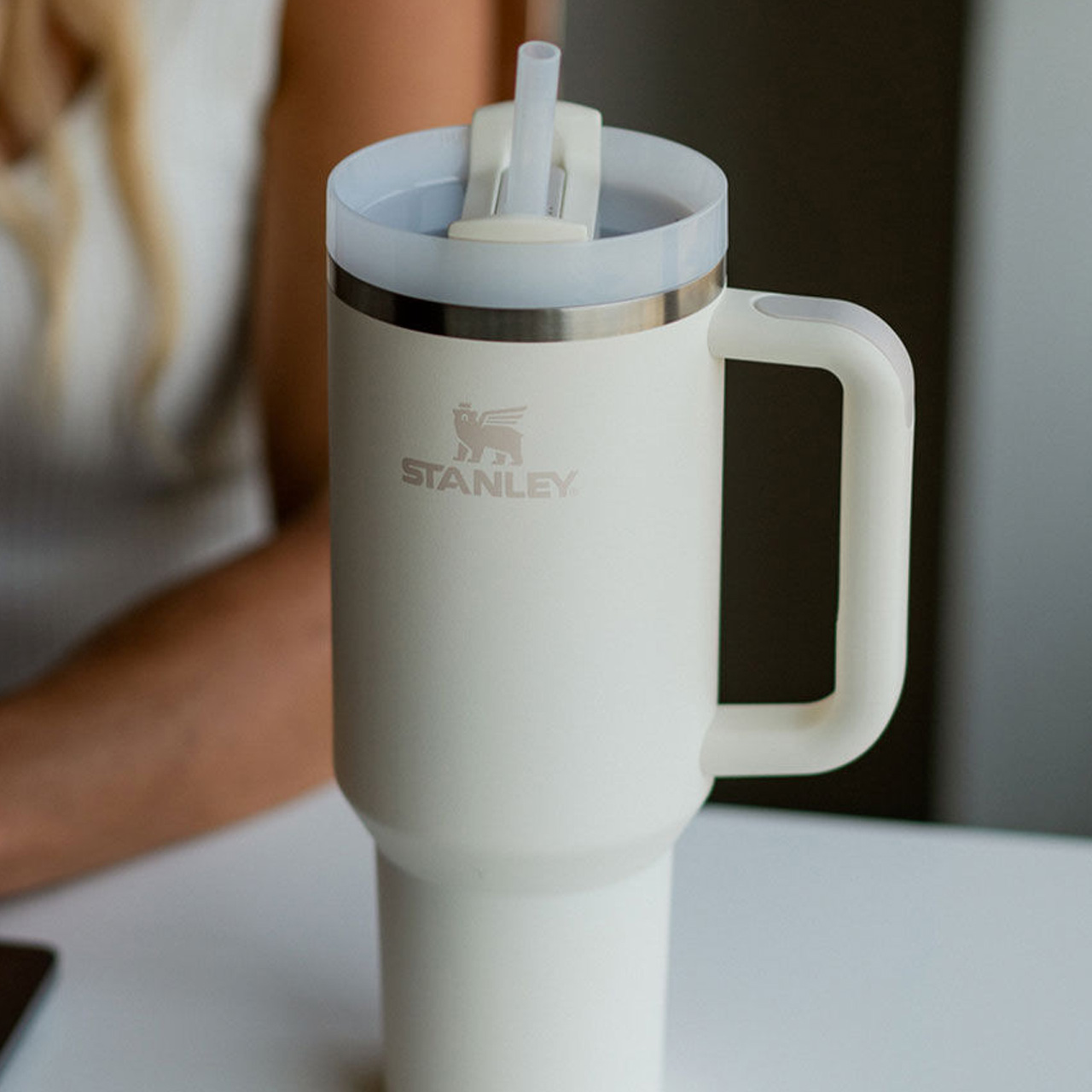 A white Stanley travel mug with a handle and straw sits on a table.