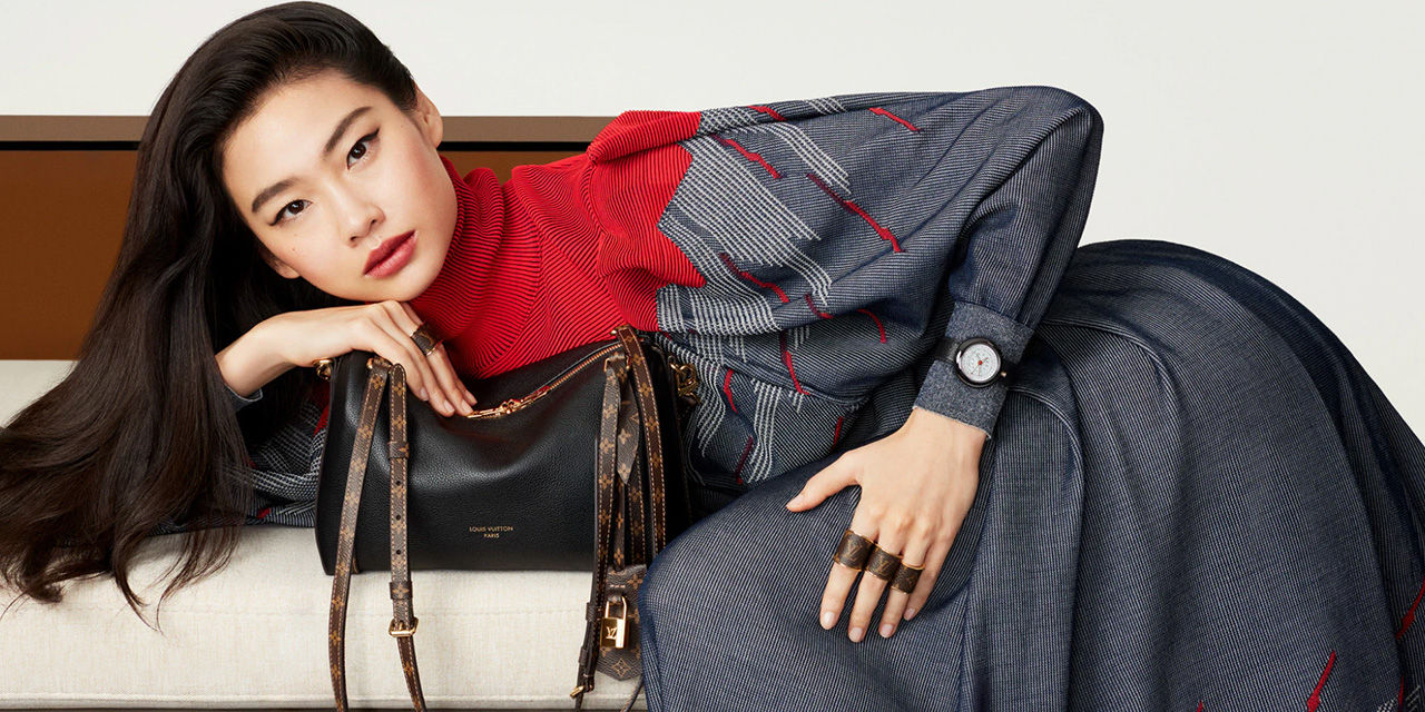 A person in a stylish outfit reclines on a sofa, holding a black handbag.