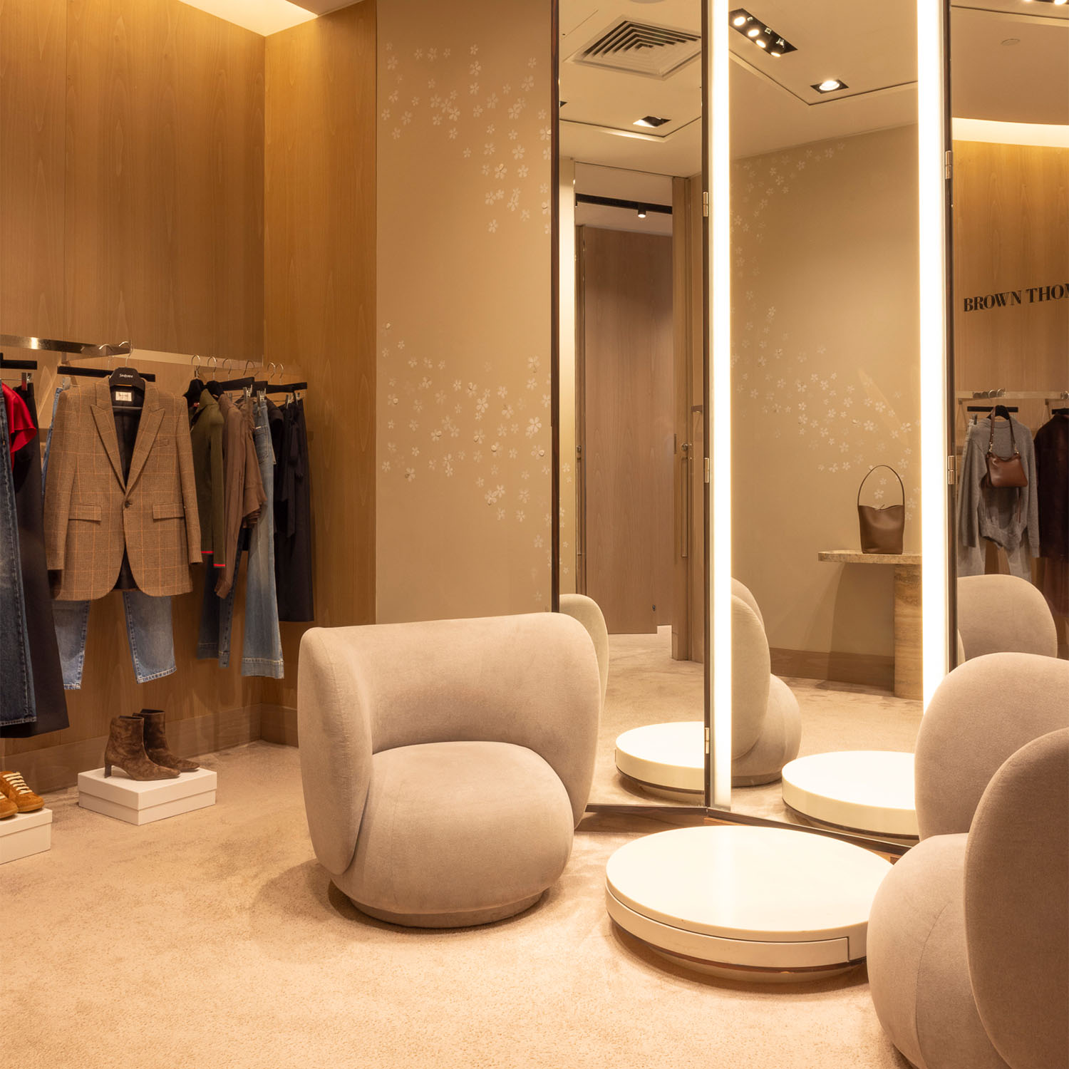 A chic fitting room with elegant gray chairs, mirrors, and clothes displayed on racks in a warmly lit, modern interior.