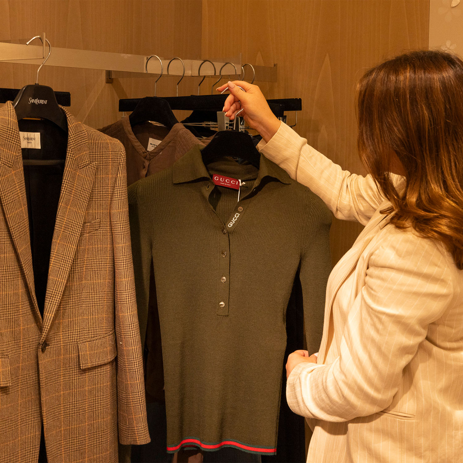 A person in a light-colored jacket selects a green Gucci top from a rack with other clothing items.