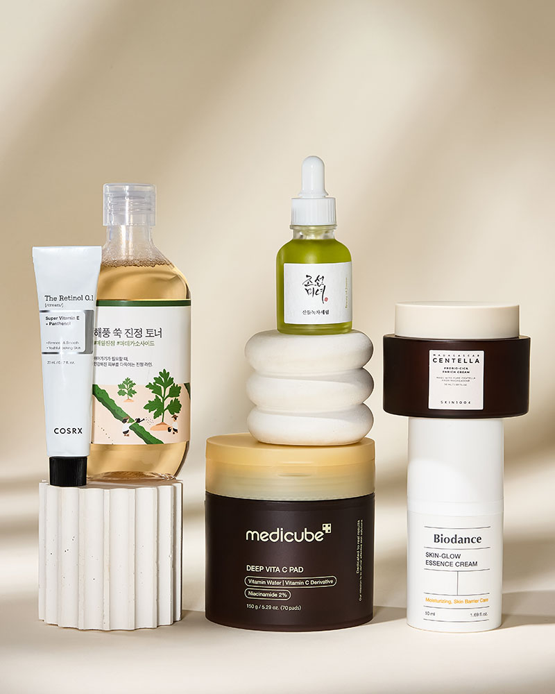 A collection of skincare products, including bottles, jars, and tubes, displayed on textured stands with a neutral background.