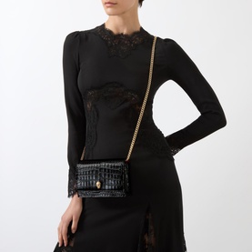 Model carrying Alexander McQueen Skull Clutch Bag in black croc-embossed leather with gold chain strap, styled with black lace-trim top and skirt