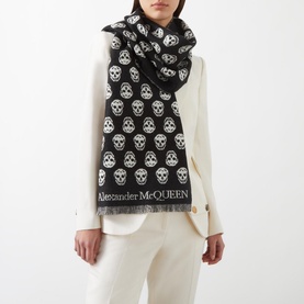 Model wearing Alexander McQueen Reversible Skull Scarf in black, featuring all-over skull print and fringe hem, styled with cream outfit