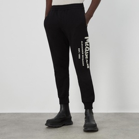 Model wearing Alexander McQueen Graffiti Logo Sweatpants in black with large white logo print, styled with black Chelsea boots