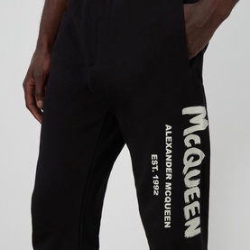 Model wearing Alexander McQueen Graffiti Logo Sweatpants in black with large off-white graffiti logo print on leg