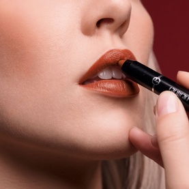 Armani Beauty Color Sketcher lip pencil in Scarlatto, shown being applied to model's lips in a warm burnt-orange-red shade