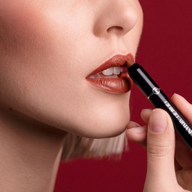 Model wearing Armani Beauty Color Sketcher lipstick in Terra, a warm brick-red shade, applied directly to lips with the slim black bullet tip visible