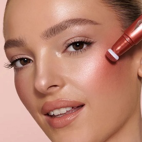 Model with Charlotte Tilbury Beauty Lightwand applied to cheek, giving a coral-pink luminous flush to skin.