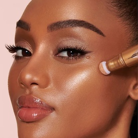 Model with Charlotte Tilbury Beauty Light Wand Highlighter in Medium/Deep applied to cheekbone, showing luminous glowing finish on deep skin tone.