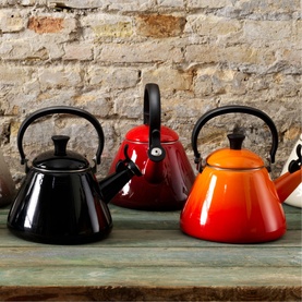 Le Creuset Kone Kettle With Fixed Whistle 1.6L in Black, shown alongside red and orange colourways on a rustic wooden shelf