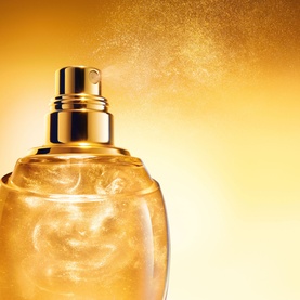 Dior J'adore Shimmering Hair Mist in gold glass bottle with spray nozzle releasing a golden shimmer mist