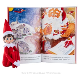 Elf on the Shelf Girl Scout Elf soft doll in red outfit with dark hair, posed beside an open illustrated Christmas storybook