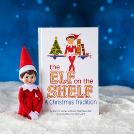 Elf on a Shelf Girl Scout Elf soft toy in red beside the Elf on the Shelf: A Christmas Tradition hardcover book, on snowy surface