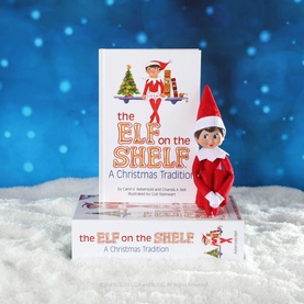 Elf on a Shelf Girl Scout Elf doll in red and white outfit posed beside the classic A Christmas Tradition storybook, on snowy display