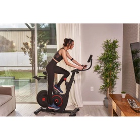 Woman riding Echelon EX5S Connect Bike in black, set up in a bright living room beside indoor plants and large windows