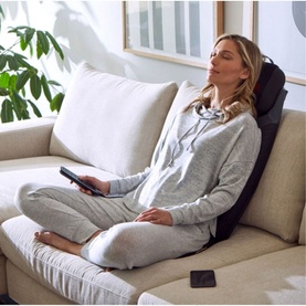 Model with Homedics 2-in-1 Shiatsu Massaging Seat Topper with Removeable Massage Pillow, seated on sofa holding remote control