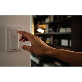 Philips Hue Dimmer Switch in white, mounted on wall, with hand pressing button, illuminated room visible in background