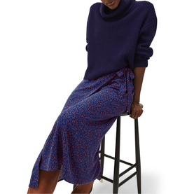Model wearing Phase Eight Karine Chunky Roll Neck Sweater in blue, styled with a blue floral midi skirt, seated on a stool