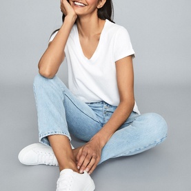 Model wearing Reiss Luana V-Neck T-Shirt in white, styled with light blue jeans and white trainers, seated on grey background