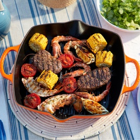 Le Creuset Signature Cast Iron Deep Square Grill 30cm in orange, filled with grilled prawns, steak, corn and tomatoes on a striped surface