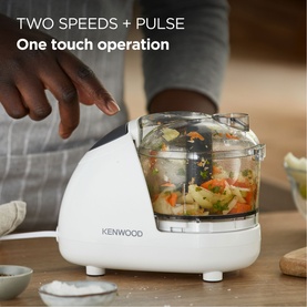 Kenwood Mini Chopper in white with transparent bowl containing chopped vegetables, hands adding ingredients above