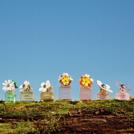 Marc Jacobs Daisy fragrance collection of six EDT bottles with floral caps displayed on moss-covered log against blue sky