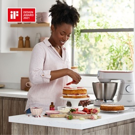 Woman decorating a layered cake using the Kenwood Titanium Chef Baker stand mixer on a bright kitchen countertop.