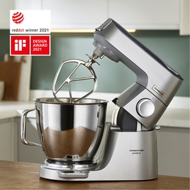 Kenwood Titanium Chef Baker XL Stand Mixer in silver with stainless steel bowl, whisk attachment, and dough hook, shown with flour.
