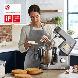 Model using a silver Kenwood stand mixer in a kitchen setting, adding butter to the bowl, with sponge cake layers on the counter.