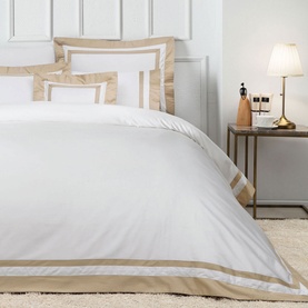 Valeron Nouveau Flat Sheet in white with tan border trim, styled on a bed with matching pillowcases and bedside lamp in background