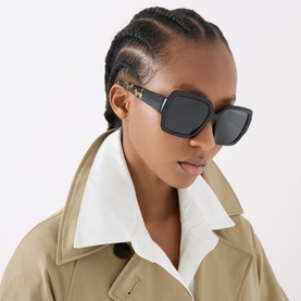 Model wearing Prada 0PR 21XS Pillow Sunglasses in black with oversized frames and tortoiseshell temple detail, styled with a beige trench coat