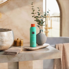 Rituals Sun Care Karma SPF30 turquoise spray bottle with orange cap, styled on marble shelf in a spa-like bathroom setting