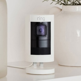 Ring Stick Up Cam Wired in white, compact indoor security camera with wide-angle lens, shown on a shelf with cable attached