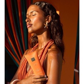 Model holding Rituals The Ritual of Karma After Sun Hydrating Lotion in an orange tube, wearing terracotta draped top and star earring.