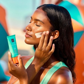 Model applying Rituals Ritual Sun Care Karma SPF50 to face, holding turquoise tube, wearing mint green top outdoors.