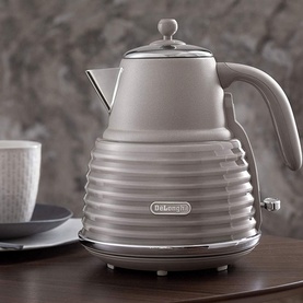DeLonghi Scolpito Kettle in beige with chrome lid and handle, ribbed body design, shown on countertop with cup in background