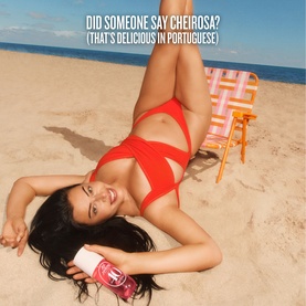 Model with Sol De Janeiro Cheirosa '40 Hair + Body Fragrance Mist, lying on sandy beach in red bikini, holding pink product bottle.