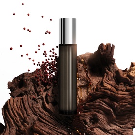 Dark brown ribbed fragrance rollerball with silver cap, displayed against textured driftwood with scattered red peppercorns