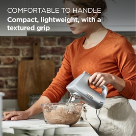 Model wearing orange top using a silver compact hand mixer to blend ingredients in a glass bowl on a kitchen counter
