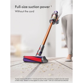 Dyson Home V10 Cyclone Absolute Cordless Stick Vacuum Cleaner in copper, shown upright on hard floor with scattered debris nearby