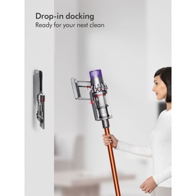 Dyson V10 Cyclone Absolute Cordless Stick Vacuum in copper, shown wall-docked on charging mount with model holding the wand