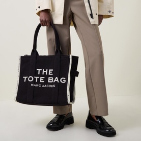 Model carrying Marc Jacobs The Jacquard Large Tote Bag in black, with white logo lettering, top handles, and boxy shape. Styled with loafers