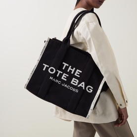 Model carrying Marc Jacobs The Jacquard Large Tote Bag in black, with white logo lettering, top handles, and boxy shape