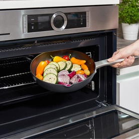 Zyliss Ultimate Pro 24cm Frying Pan with Spout in stainless steel, filled with fresh vegetables, being placed into a stainless steel oven