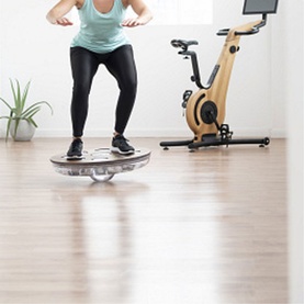 Model wearing black leggings using NOHrD Eau-Me Balance Board in light brown ash wood, water-filled design, in a home gym setting