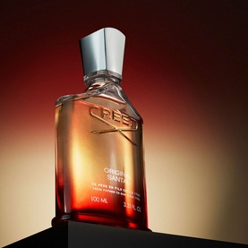CREED Original Santal Eau de Parfum in amber glass bottle with silver cap, displayed on dark plinth against warm red-gold background