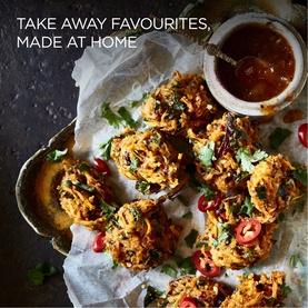Homemade onion bhajis garnished with fresh coriander and chilli, served on parchment with a dipping sauce, styled on a dark surface.