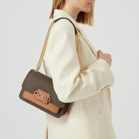 Model carrying MMK Heather Large Logo-Canvas Shoulder Bag in brown with gold chain strap and logo plaque, styled with cream blazer